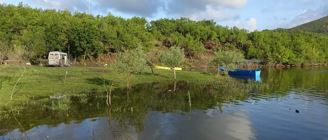 agora camping, agora farmhouse camping, Campingplatz in Albanien, großer See mit zwei bunten Booten, dahinter eine Wiese mit einem Wohnmobil und im Hintergrund, Wald